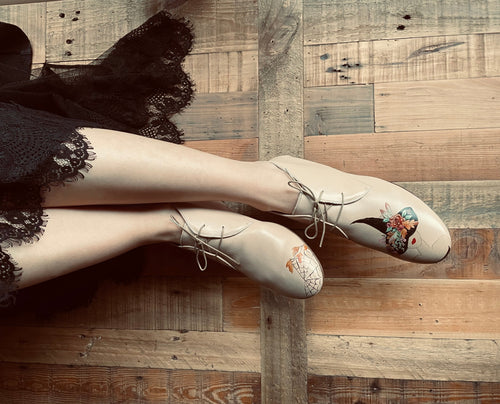 A pair of beige women's flats with decorative elements on the straps, displayed against a wooden backdrop with a person's lower legs and a black lace dress in the background.
